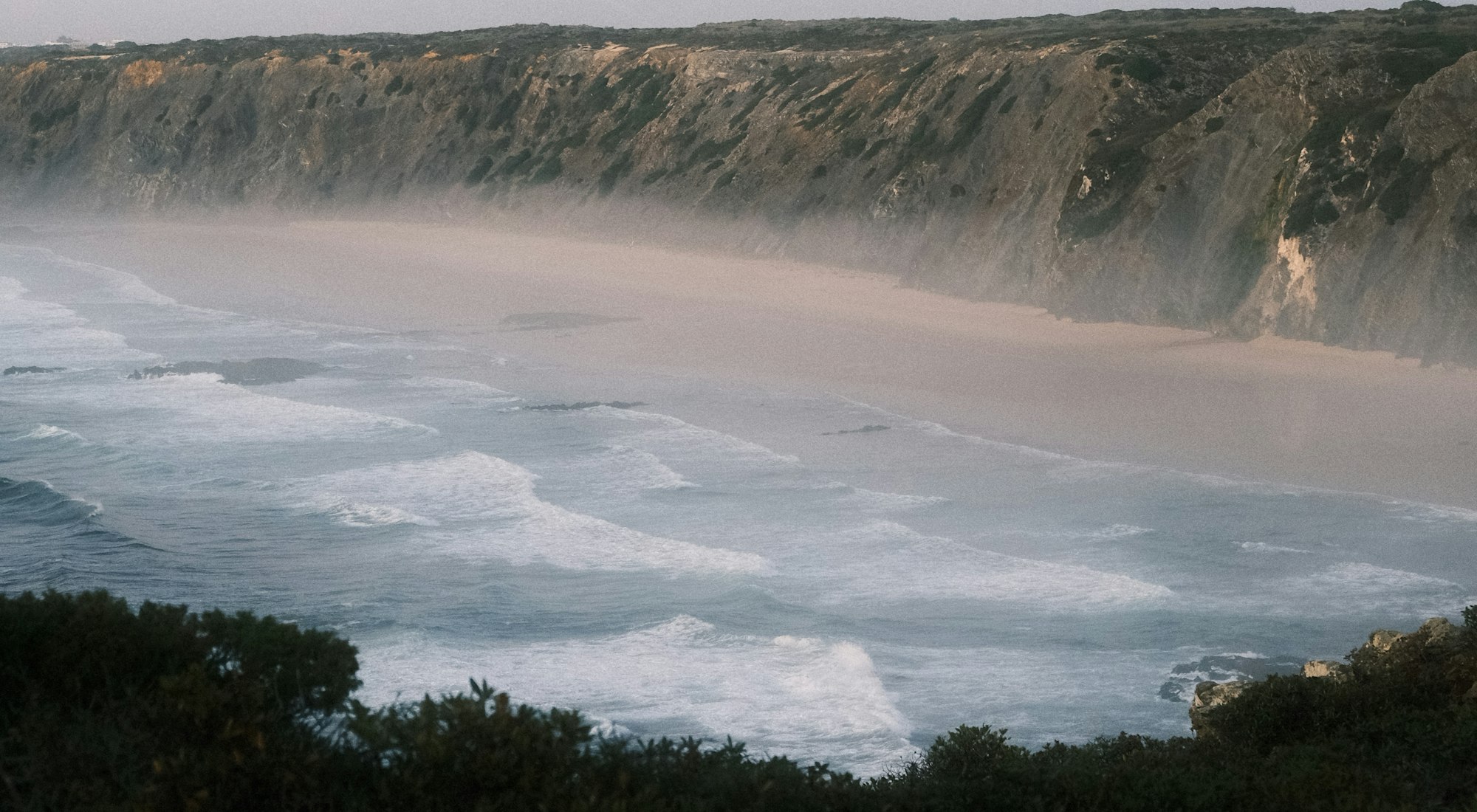 Waves rolling into a wide coastal cove below misty cliffs